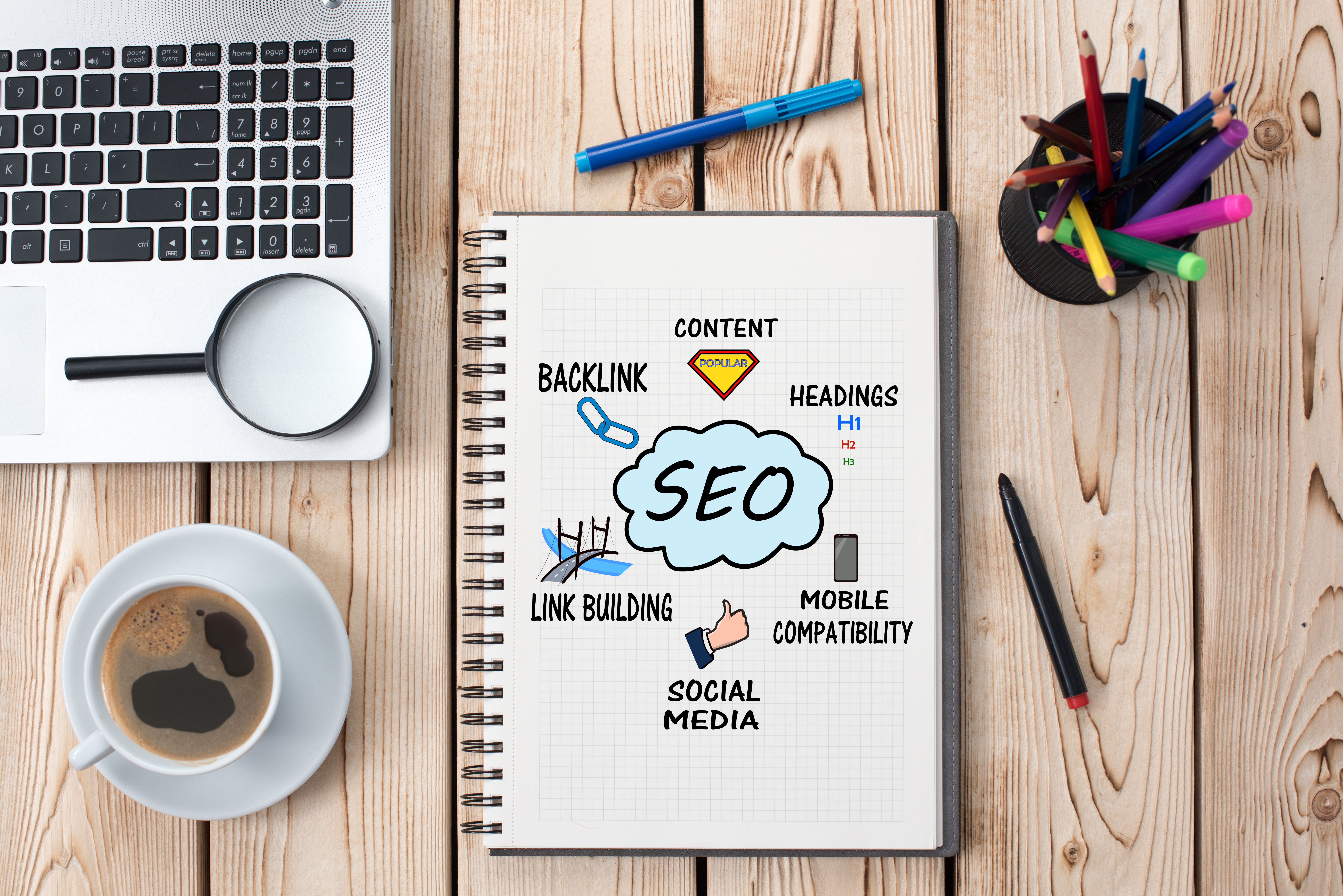 a notebook depicting elements of SEO on a wooden table with a laptop, magnifying glass, cup of coffee and pens